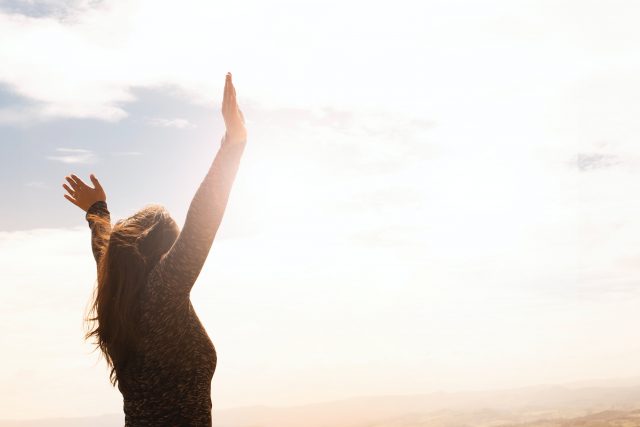 woman raises her hands towards a bright sunny sky. Photo by Daniel Reche